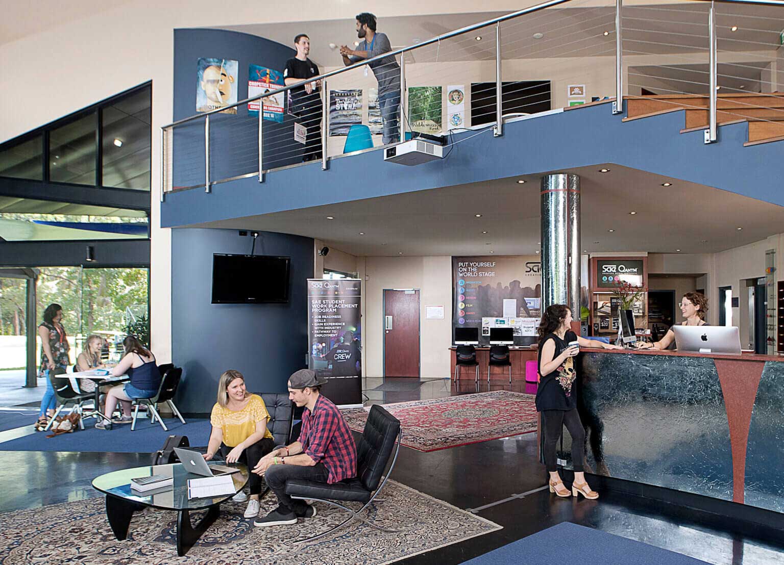 SAE Byron Bay campus foyer. Mezzanine, front desk and tables and chairs all feature people talking and chatting.