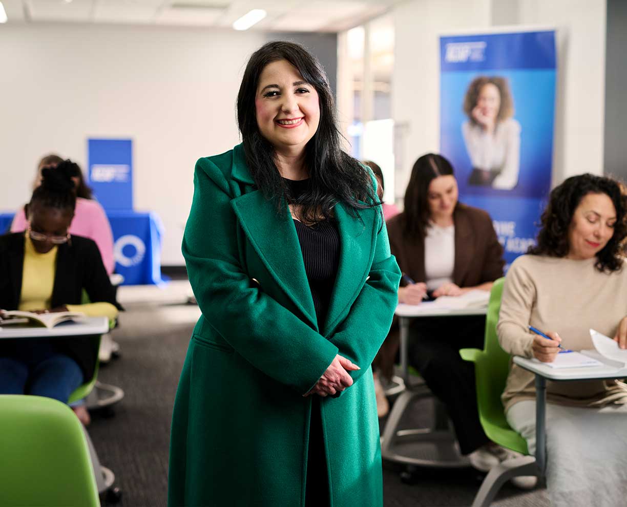 Woman with dark hair wears a dark green wool coat. People sit at individual desks looking down at table (as if in a classroom).