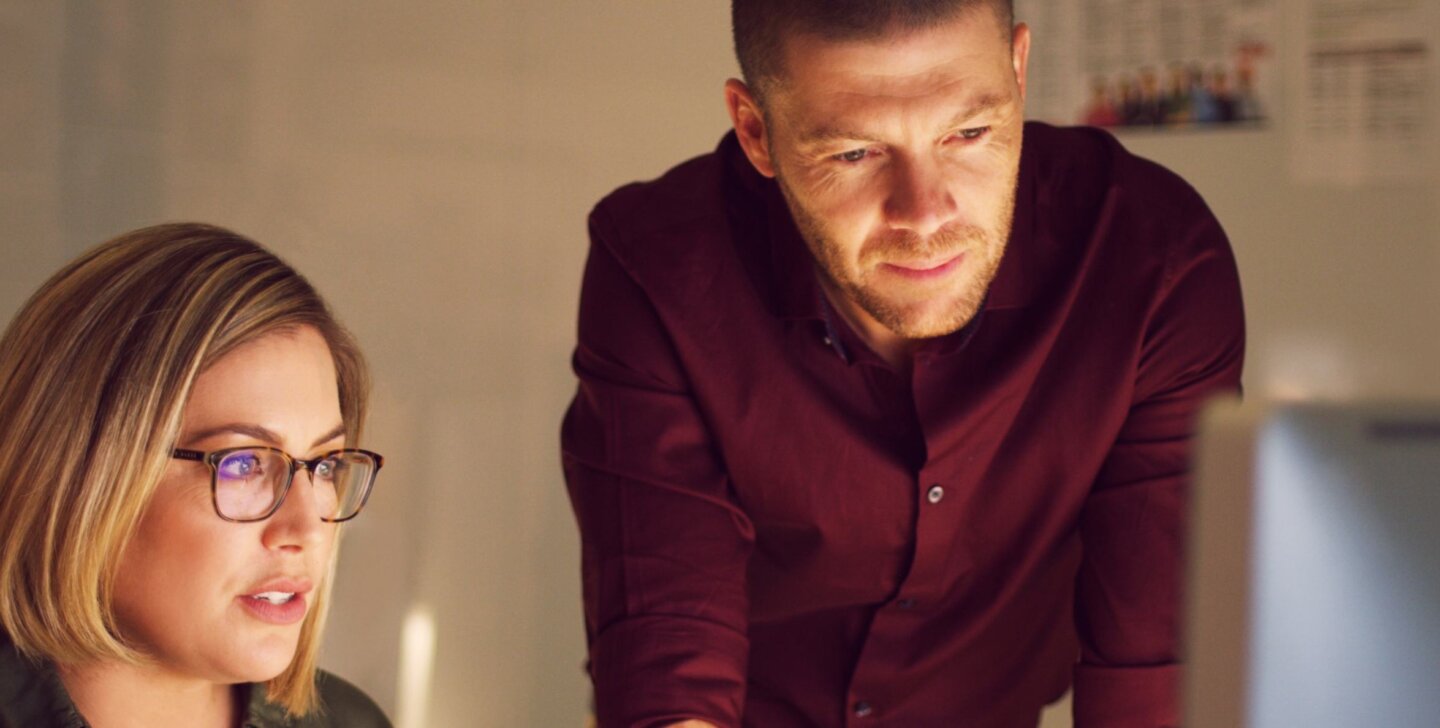 A woman with glasses sitting at, and man standing over a desk looking at a desktop monitor