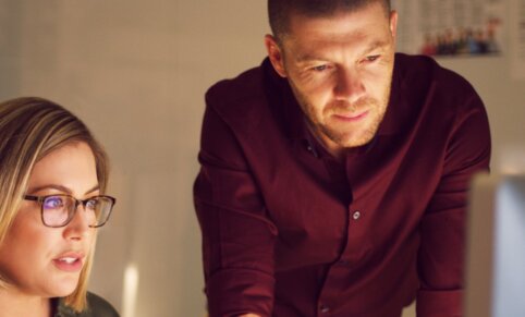 A woman with glasses sitting at, and man standing over a desk looking at a desktop monitor