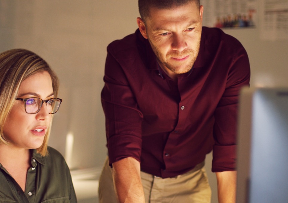 A woman sitting and man standing at a desk, looking towards a laptop screen thumbnail image