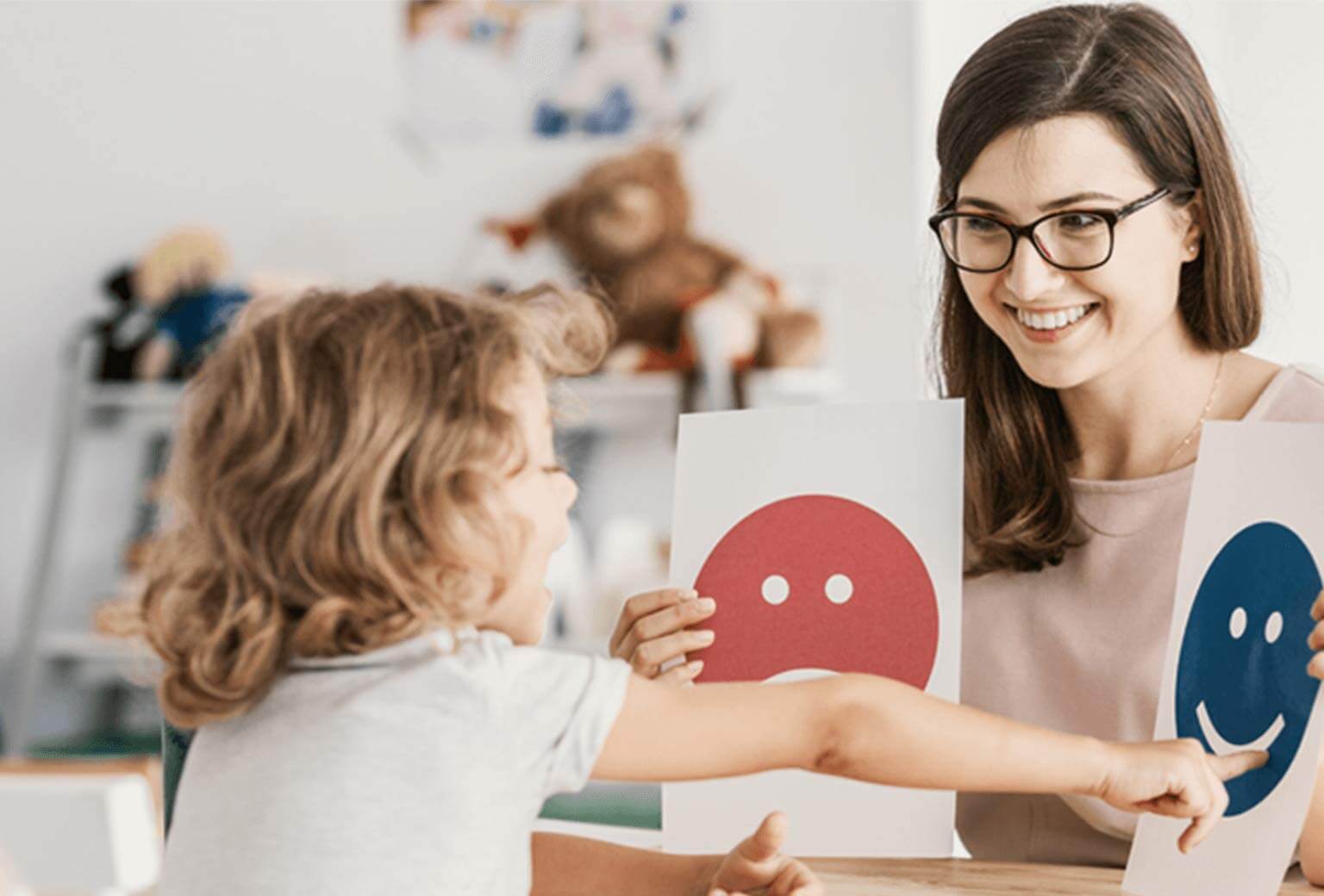 Woman sits at desk holding two paper cards with an image on each. A young child sits in front of her and is pointing at one and speaking.