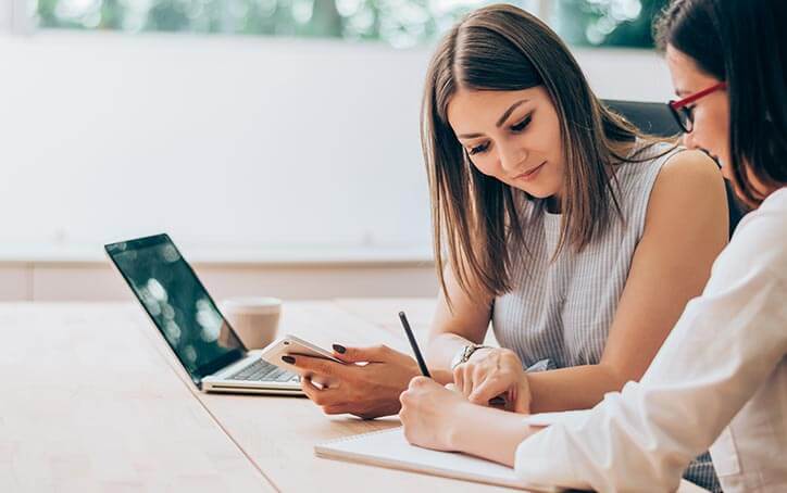 Woman leans over writing pad. She holds a phone and points to page that the other woman is writing on. A laptop and coffee sit on desk in front of woman.