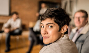 Woman with short cropped hair looks over her shoulder to camera. The background features group of people in circle of chairs.