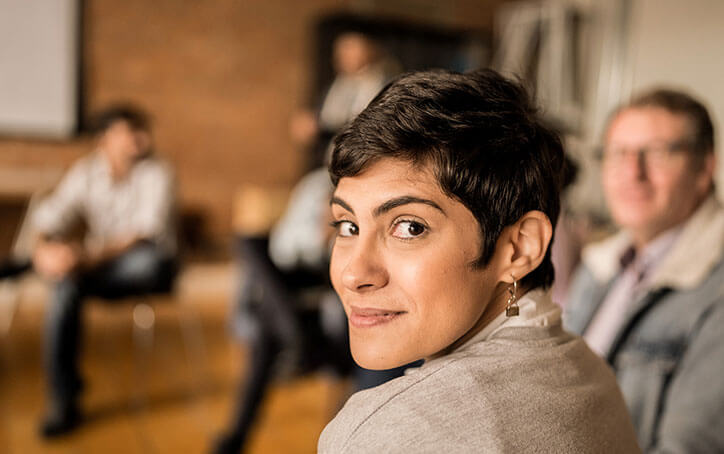 Woman with short cropped hair looks over her shoulder to camera. The background features group of people in circle of chairs.