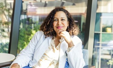 Woman sitting in chair in front of window. Leaning on arm rest, hand under chin. She smiles at camera.