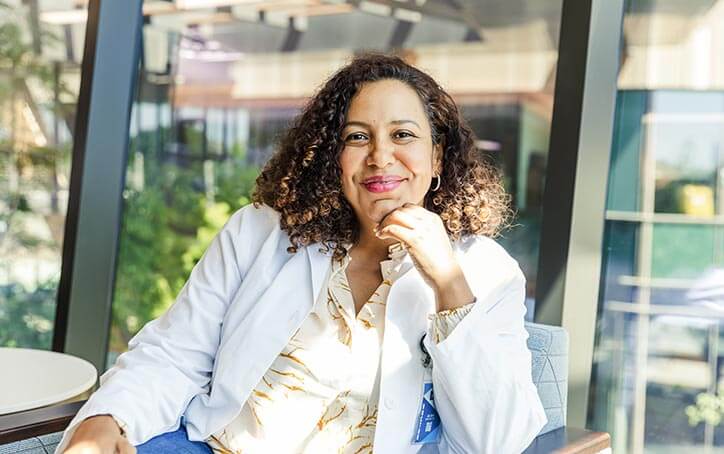 Woman sitting in chair in front of window. Leaning on arm rest, hand under chin. She smiles at camera.