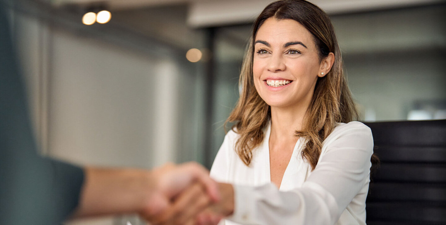 Woman smiles and shakes hand of person off camera. She holds a document and sits at a desk.