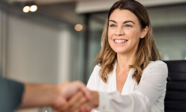 Woman smiles and shakes hand of person off camera. She holds a document and sits at a desk.