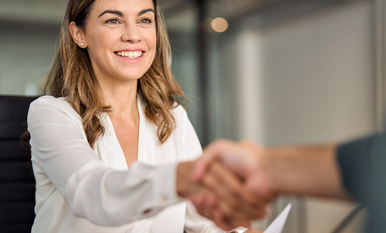 Woman smiles and shakes hand of person off camera. She holds a document and sits at a desk.