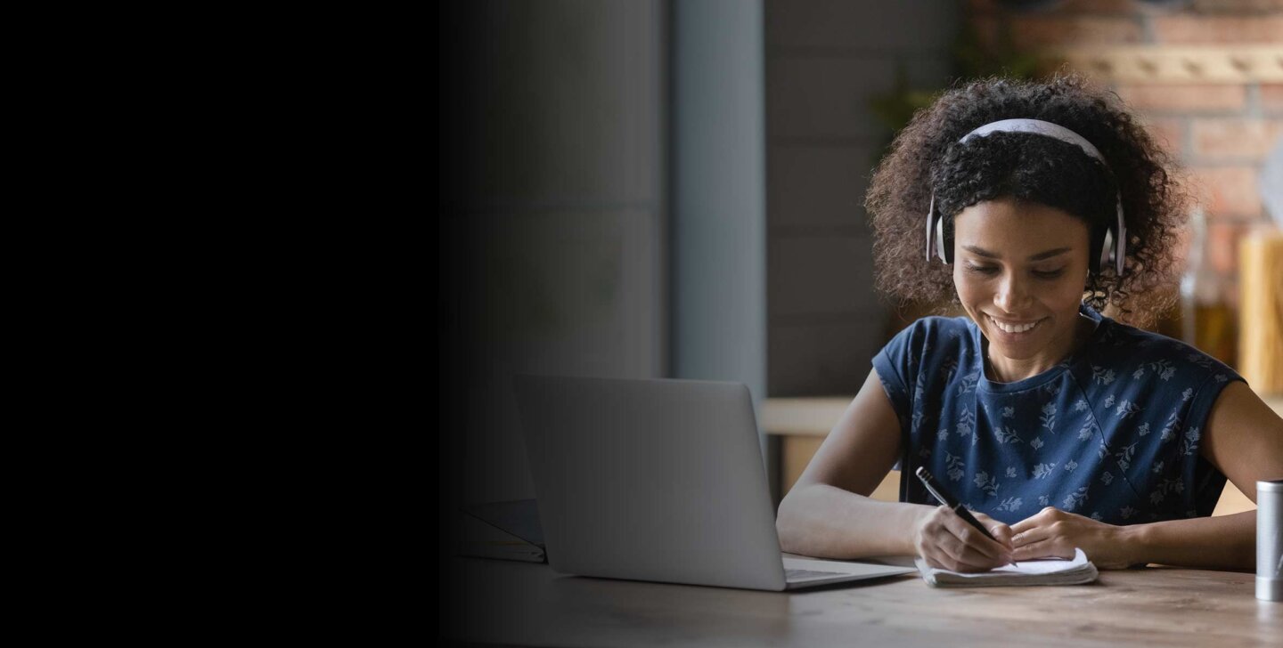 Curly haired woman wears wireless headphones and looks down at notepad she is writing on with a pen. She smiles. A laptop and glass of water on desk to the side and front of her. She appears to be in a kitchen - working at the table. A heat mat and food jars of pasta blurry in background.