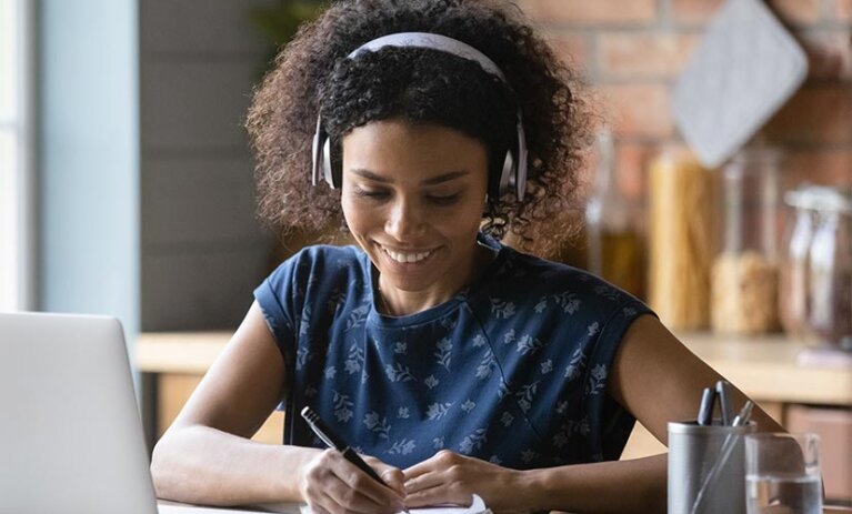 Woman with curly hair wears headphones and looks down at notepad with pen in hand. She is smiling. A laptop, pen holder and glass of water on desk in front of her. She seems to be in a kitchen - food jars of pasta blurry in background.