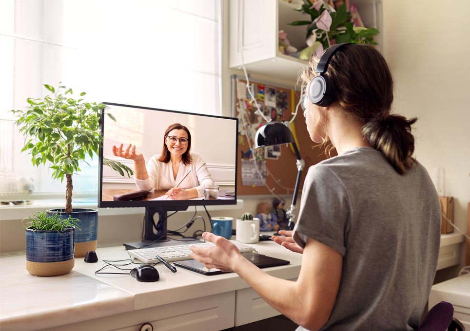 Woman sits in front of computer monitor with headphones on. She is sitting in an home office and the monitor displays an image of someone speaking to camera. Like a Zoom or virtual video call.