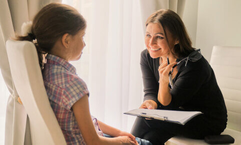 A woman sitting on a chair with a clipboard in one hand and a pen and reading glasses in the other hand, leaning towards a young child sitting in a chair opposite her | Bachelor of Social Work