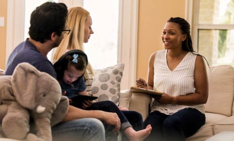 A couple sits on a couch. A young child on a tablet is on the mans lap. A woman sits opposite on another couch. She is speaking and smiling and holds a clipboard and pen in her hand.