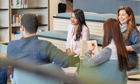 A group of people sit around a table - lounge area. Bookshelves and books in background.