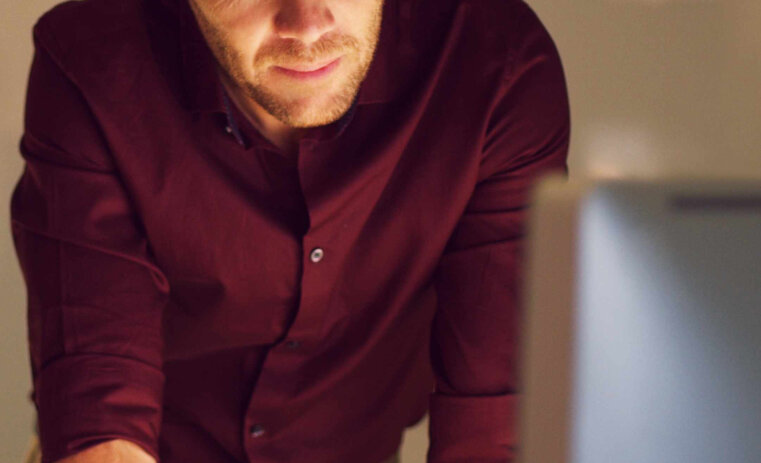 A man standing with his arms on a desk, looking towards a laptop screen