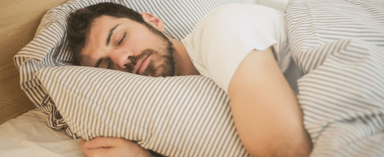 Man with a beard and wearing a white tee shirt - rests with head on pillow and doona covering him. He is lying in bed and the bedhead is just visible in background.