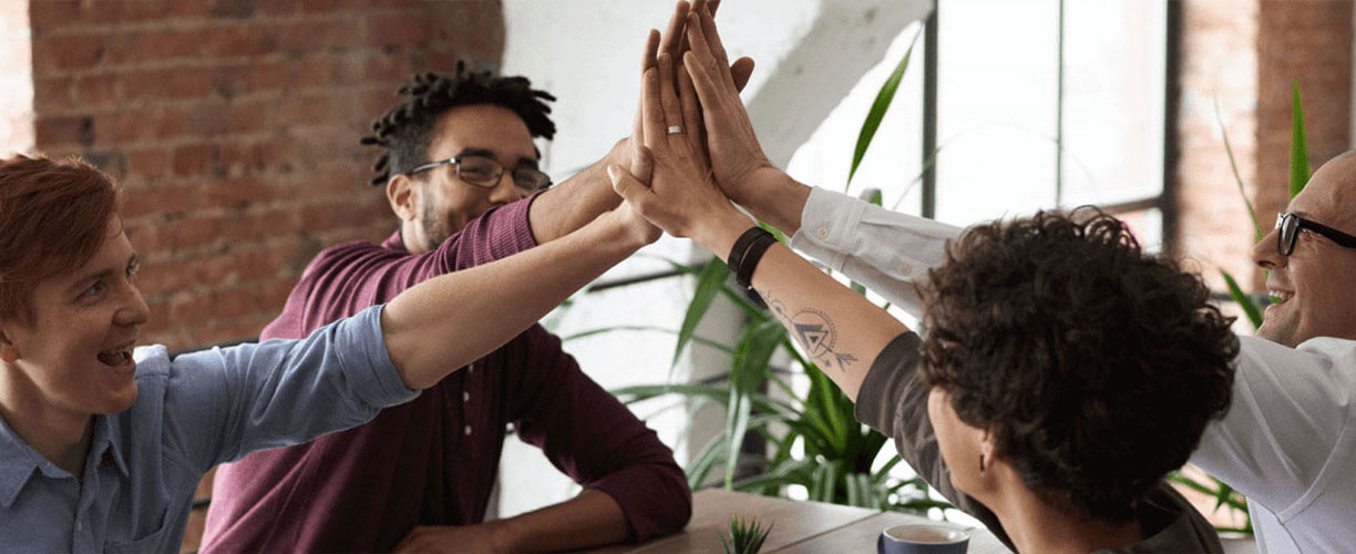 Group of four people sit around a table. Each one are raising one arm - there hands meet in the centre of table - high in the air. High five pose with four work colleagues.