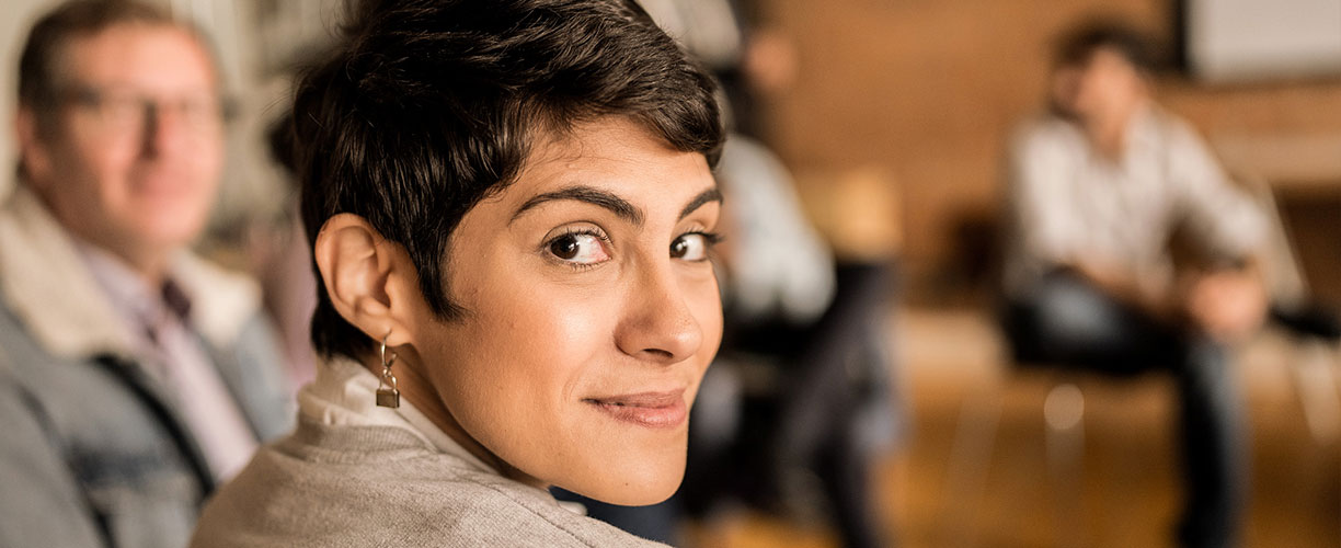 Woman with cropped hair looks over her shoulder and at camera. Group of people in background appear to be sitting in a circle on chairs.