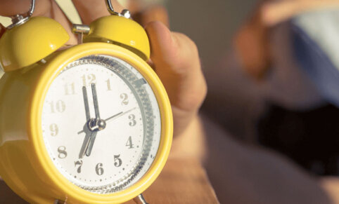A yellow metal clock with two alarm bells atop reads 7. Bright light and shadows are seen on wall behind clock. A persons hand is resting on top of alarm bells.
