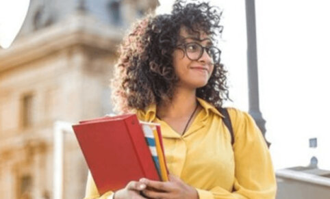 Woman standing in front of a building. Curly hair, glasses and holding a red binder and textbooks.