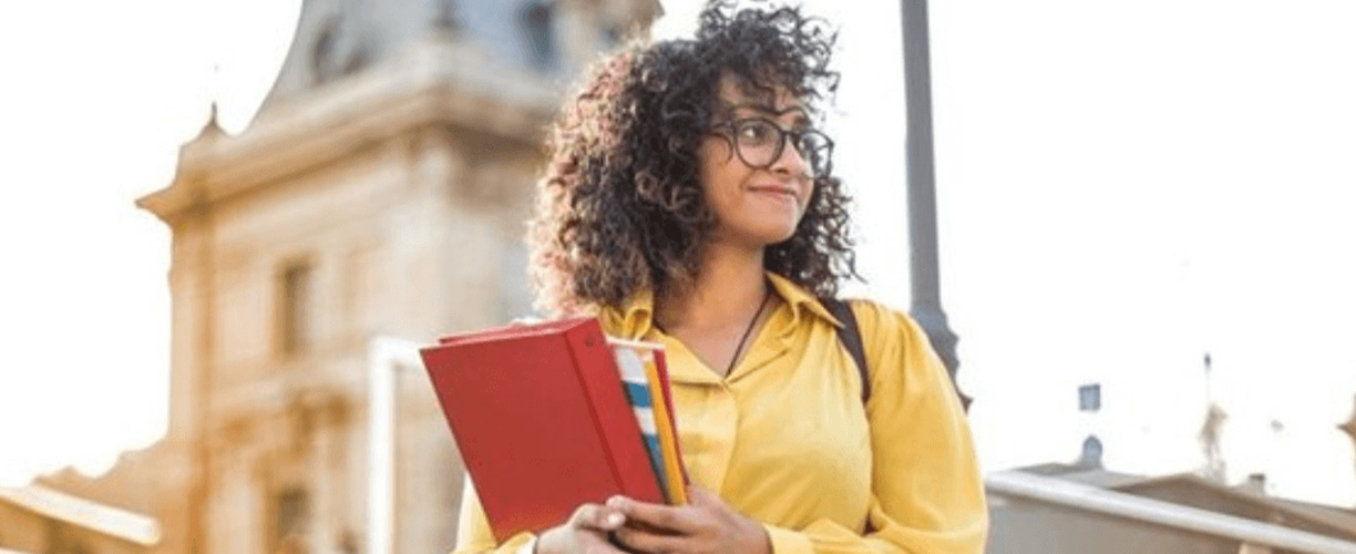 Woman standing in front of a building. Curly hair, glasses and holding a red binder and textbooks.