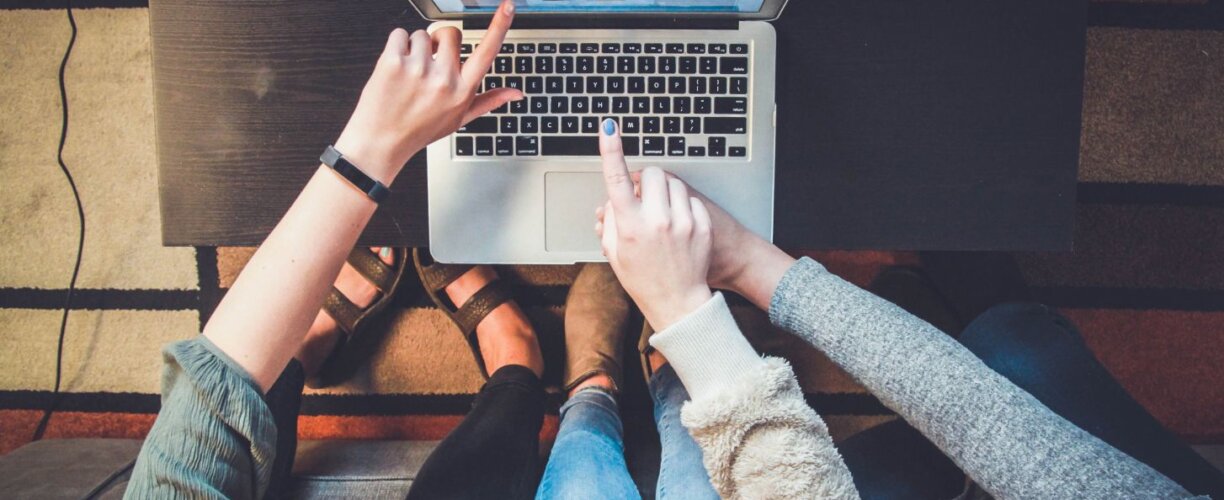 Aerial view of three students sitting and pointing towards a laptop screen, with only their arms and hands in view