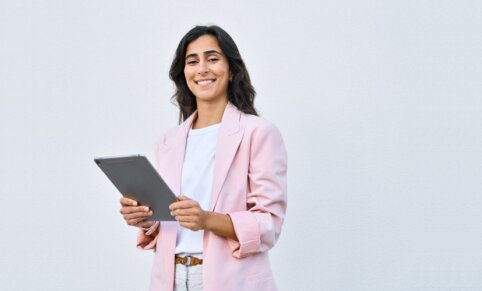 A woman wearing a pink suit jacket, standing with a tablet in hand in front of a grey background