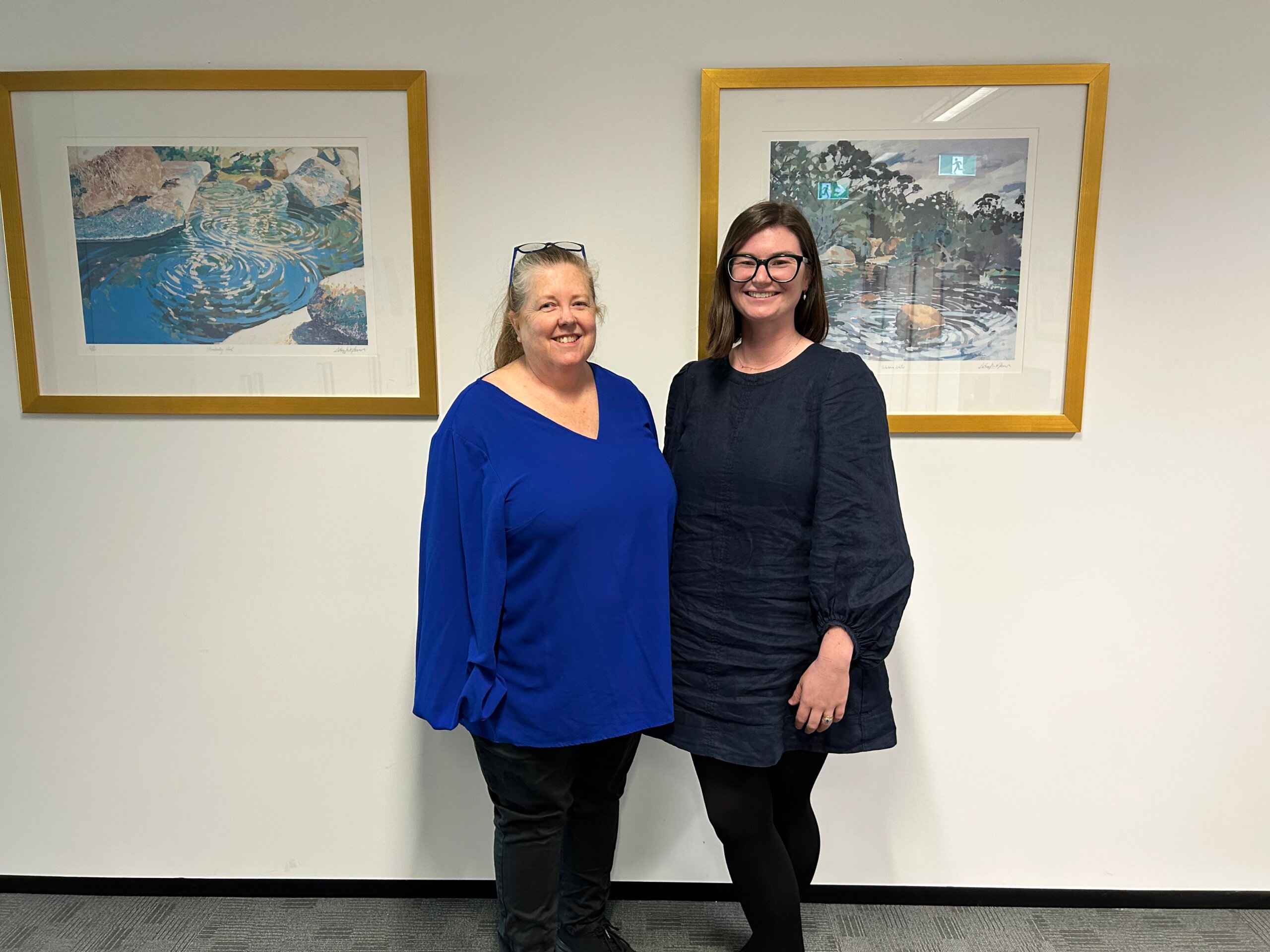 Two women one in a blue top and blank pants and another in a grey dress pose side by side in a corporate office