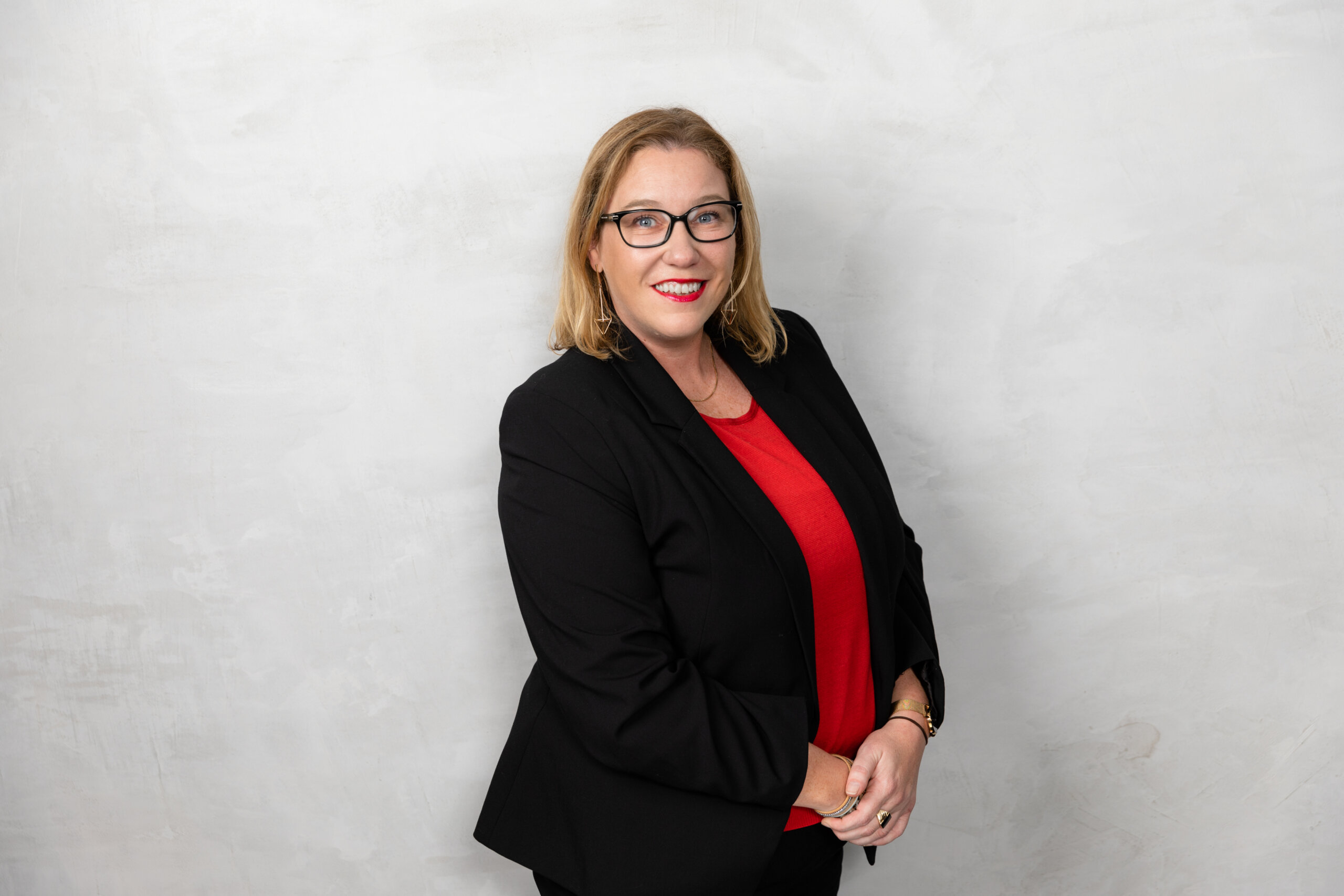 Woman with blonde hair and glasses in corporate attire smiles in front of grey background