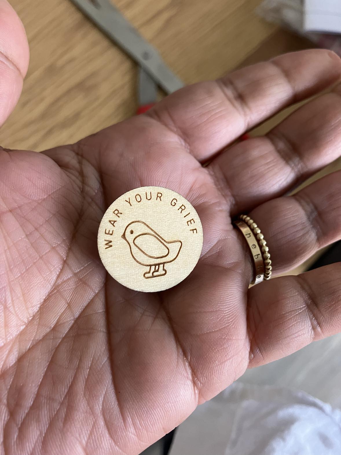 hand holding a wooden lapel pin that says 'wear your grief' with a picture of a small bird