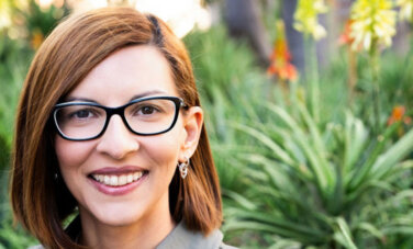 woman with red hair and glasses smiles in front of a background of greenery