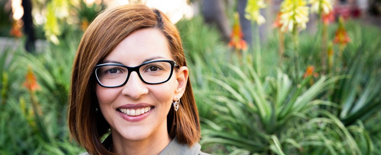 woman with red hair and glasses smiles in front of a background of greenery