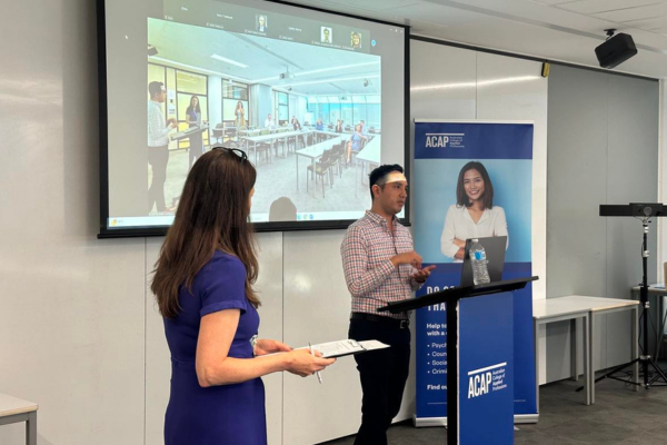 woman in blue dress looks towards young man in button up shirt giving a presentation with powerpoint