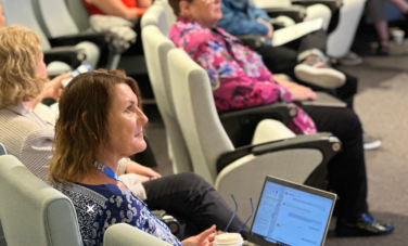 woman sits in lecture theatre filled with people. she has a coffee next to her. Laptop on her lap. And has removed her reading glasses.