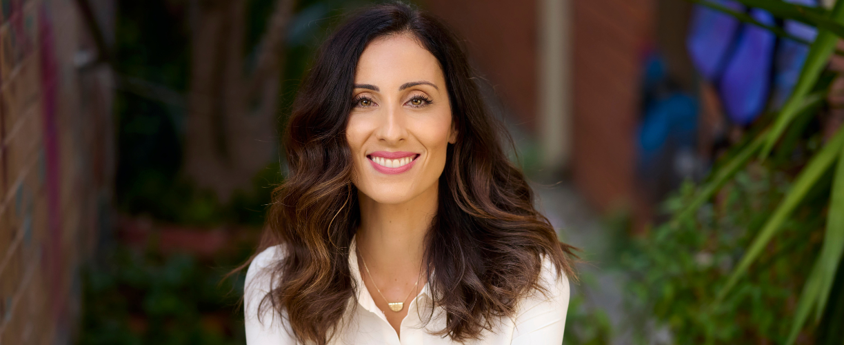 Woman with long brown hair and pink lipstick in a white collared shirt smiles at camera