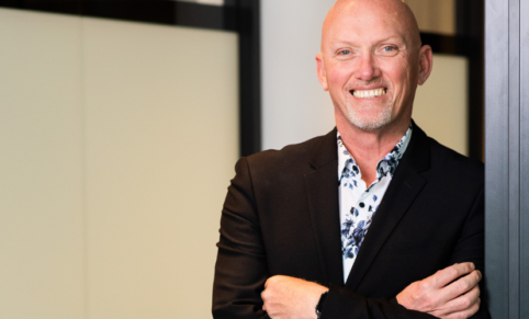 Man with bald head and a big friendly smile crossed arms in a shirt and suit jacket in front of a corporate office background