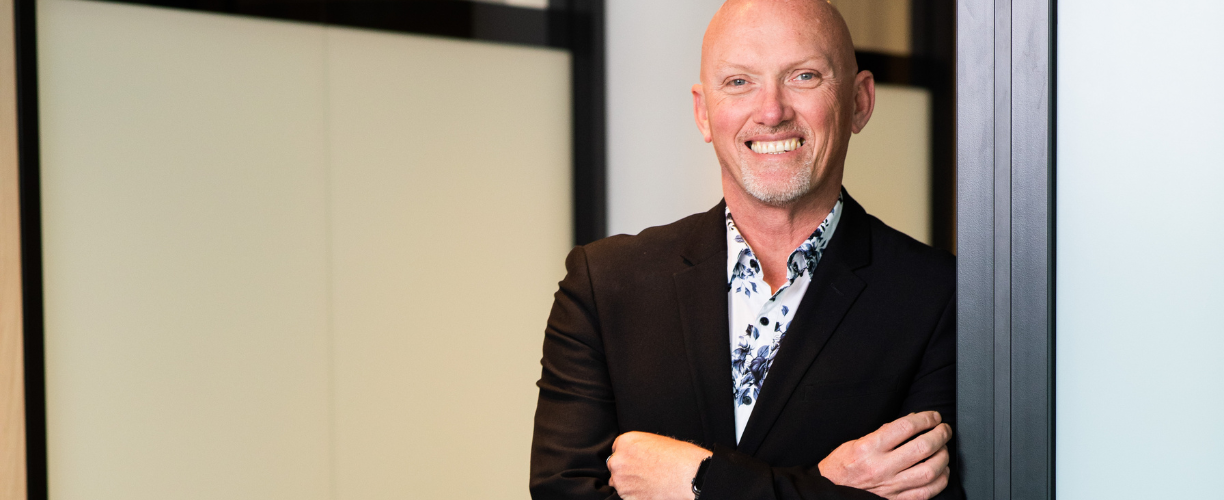 Man with bald head and a big friendly smile crossed arms in a shirt and suit jacket in front of a corporate office background