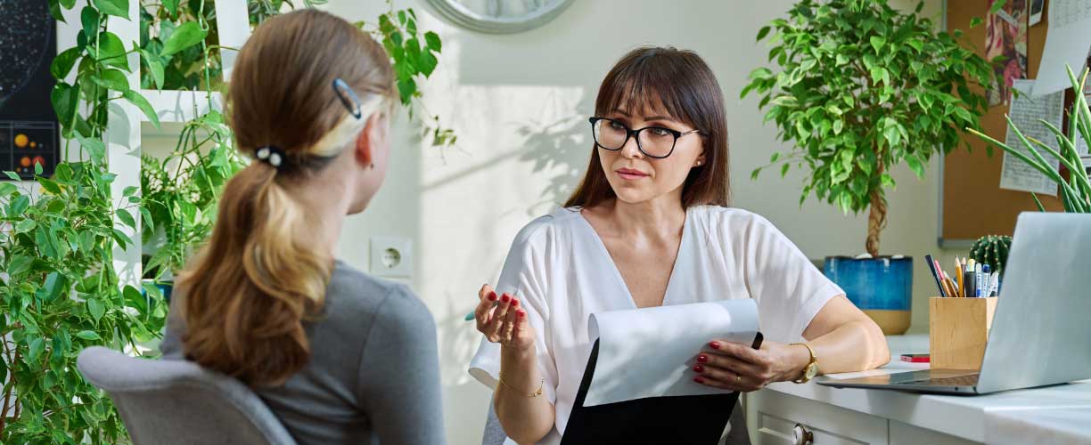 woman in office setting - sitting at desk with laptop on table. She holds a clipboard and pen in hand while speaking to a client | psychology career. A young person is seen in foreground, back to camera sitting in chair facing speaker