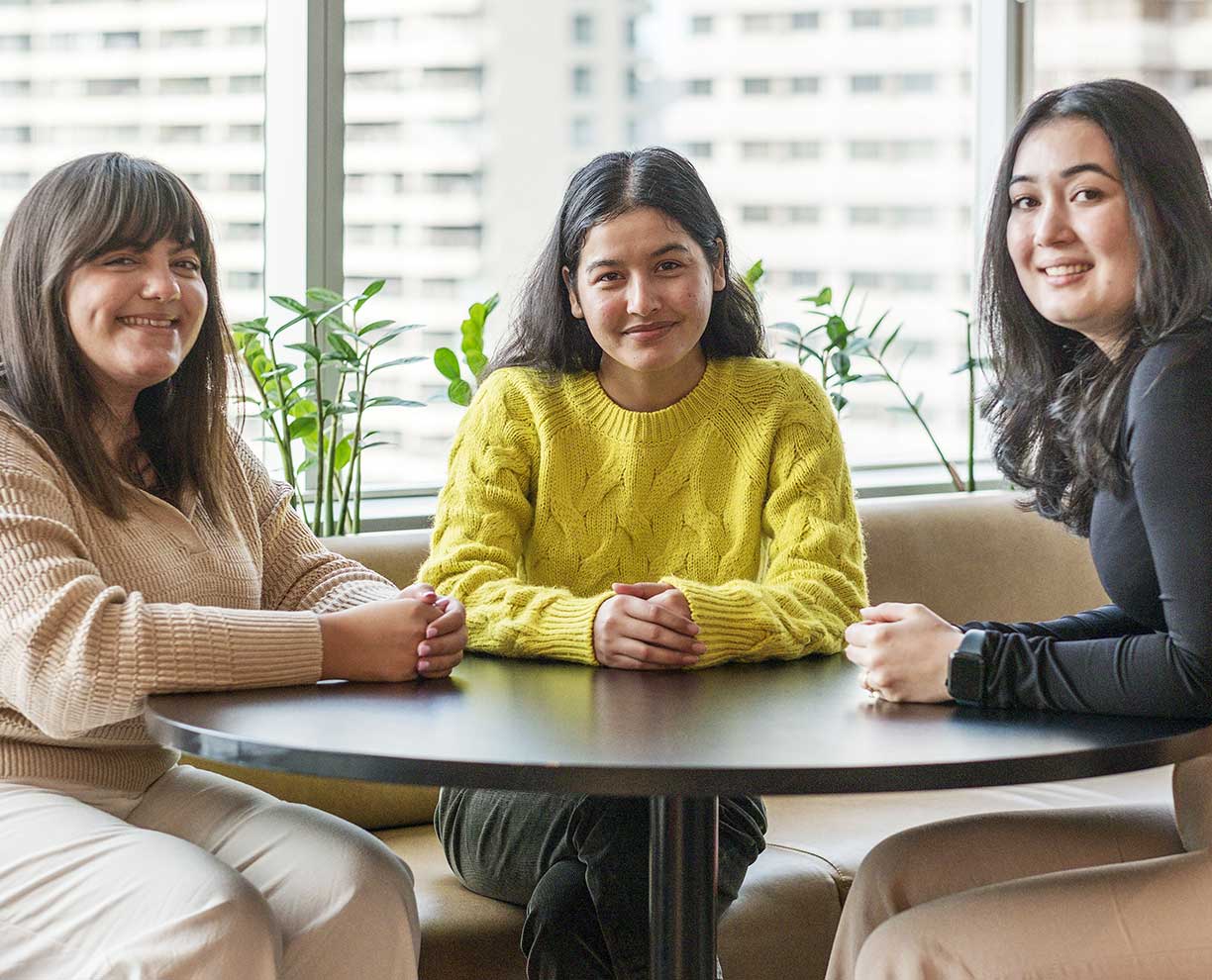 Three dark haired young women sit around a round table. Hands clasped on table in front of them.