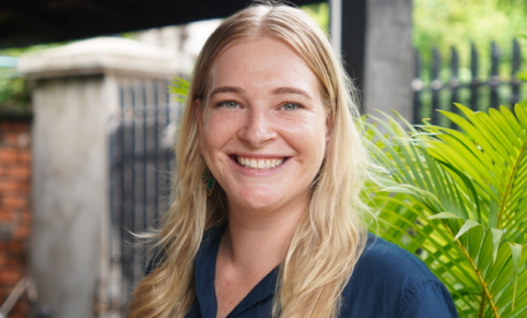 Woman with blonde hair smiles into camera in front of a background with palm leaves and a motorcycle