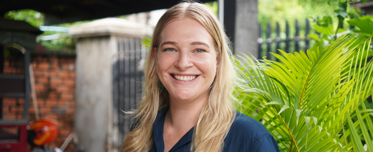 Woman with blonde hair smiles into camera in front of a background with palm leaves and a motorcycle