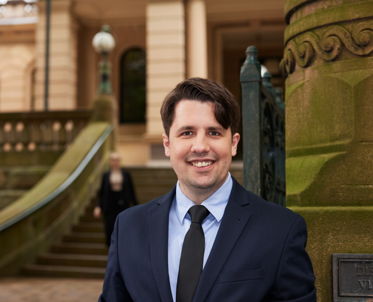 Profile photo of Anthony, Graduate Diploma of Legal Practice student standing outside a courthouse wearing a navy blue suit and smiling towards camera