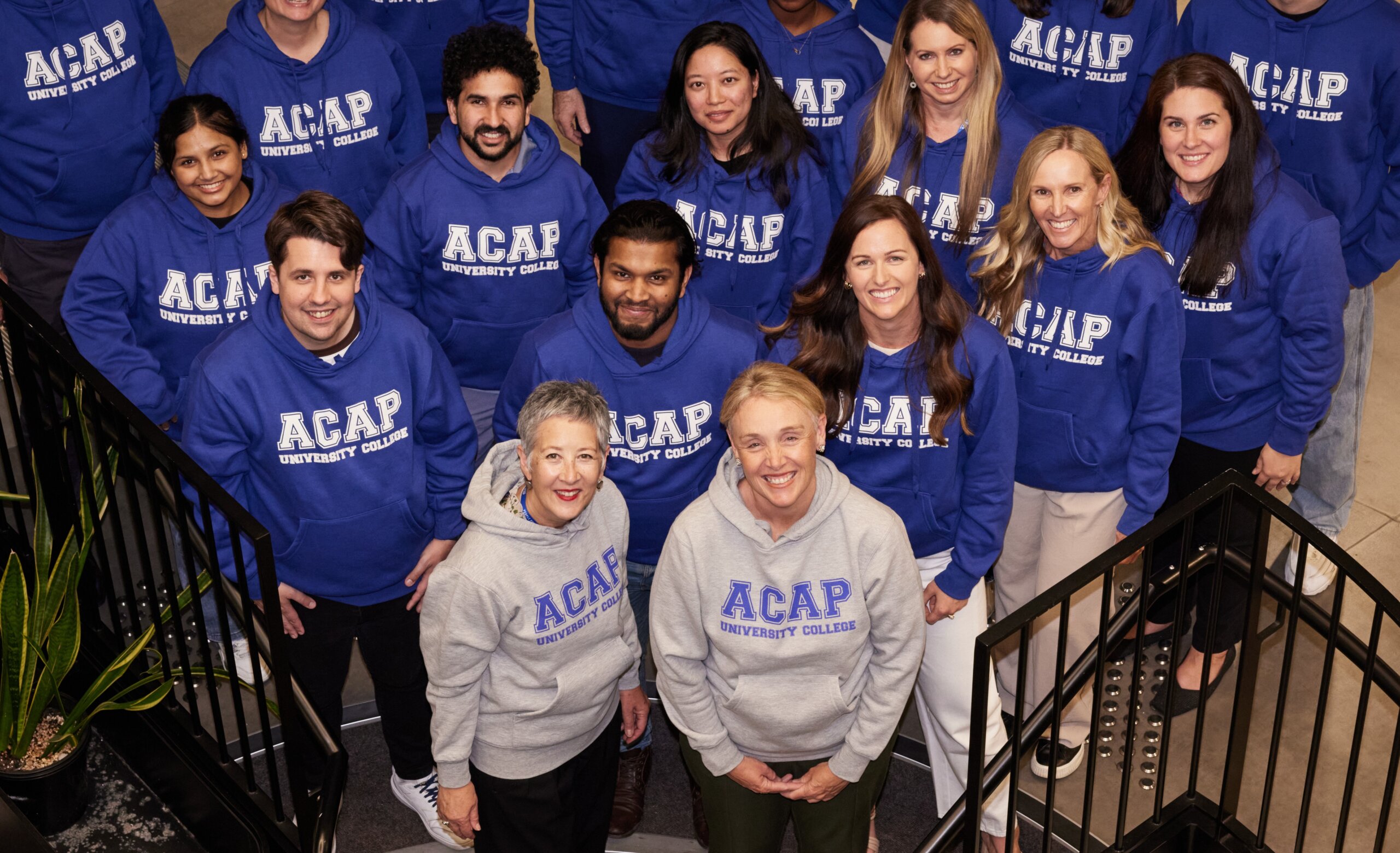 A group of people wearing hoodies with 'ACAP Universtiy College' printed on them look up at the camera smiling